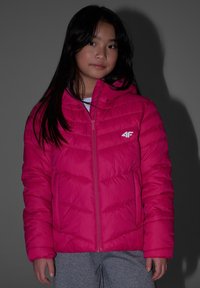 Jeune personne aux longs cheveux foncés portant une veste matelassée rose vif et un pantalon gris, debout devant un fond gris.