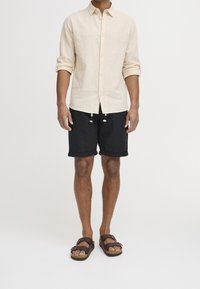 Lichtbeige linnen shirt, zwarte shorts met opgerolde manchetten, bruine sandalen met gespen. Minimalistisch ontwerp, casual uitstraling.