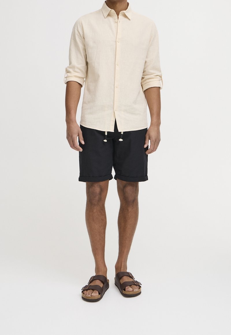 Lichtbeige linnen shirt, zwarte shorts met opgerolde manchetten, bruine sandalen met gespen. Minimalistisch ontwerp, casual uitstraling.