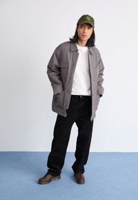 Chaqueta gris de gran tamaño con dos bolsillos frontales, camiseta blanca, pantalones negros, zapatos marrones de cordones y una gorra verde con logotipo.