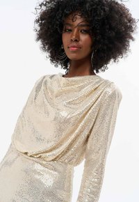 Robe dorée scintillante avec un décolleté drapé, des manches longues et une silhouette ajustée. Recouverte de sequins pour une texture réfléchissante.