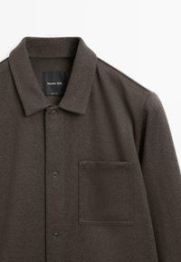 Bruine, geknoopte shirt gemaakt van zachte stof, met een enkel borstzakje en twee drukknoopjes aan de voorkant. Etiket zichtbaar binnenin de kraag.