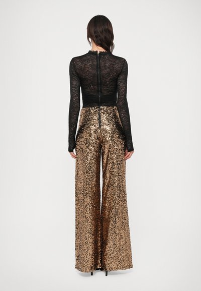 Must mustriga pikkade varrukatega topp mustast pitsist, millel on laineline kaelus, koos laia säärega kuldsete sädelevate pükstega, millel on tagumine lukku sulgumine.