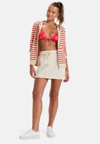Bikini rojo con lunares, con un lazo en la parte delantera, acompañado de una falda corta beige con cordón y una sudadera con cremallera a rayas en rojo y crema. Zapatillas blancas.