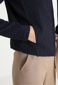 Námořnická strukturovaná bunda s jemným leskem, s přiléhavým střihem a bočními kapsami, nošená přes světle béžové kalhoty s šitími detaily.