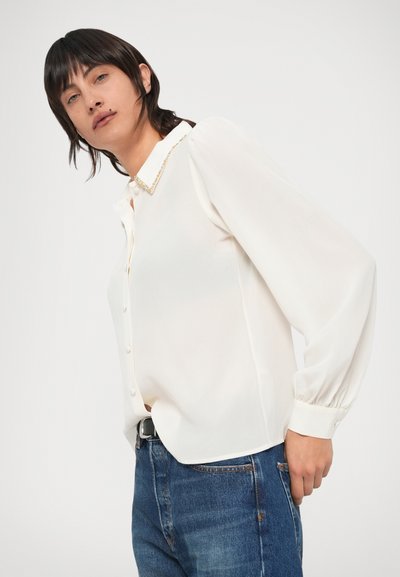 Λευκό πουκάμισο με γιακά και διακοσμητικό με χάντρες, μακριά μανίκια και κουμπιά, συνδυασμένο με μπλε τζιν παντελόνι.