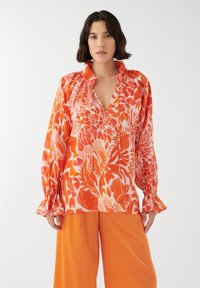 Blusa a maniche lunghe arancione con motivo floreale bianco, dotata di scollatura a V e dettagli arricciati, indossata con pantaloni arancioni a gamba larga abbinati.