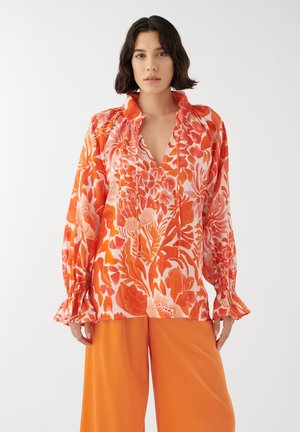 Blusa a maniche lunghe arancione con motivo floreale bianco, dotata di scollatura a V e dettagli arricciati, indossata con pantaloni arancioni a gamba larga abbinati.
