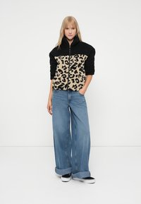 Kvinne står med hendene i lommene iført svart og beige leopardmønstret fleecejakke, brede blå jeans og svarte joggesko.