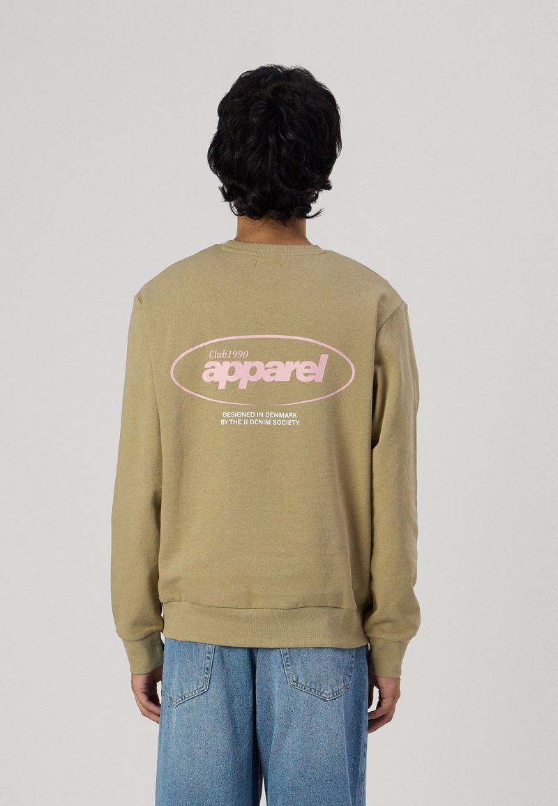 Felpa color khaki con testo grafico rosa sul retro, presenta polsini e orlo a coste, vestibilità comoda. Abbinata a jeans blu chiaro.