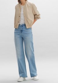 Veste bomber beige, T-shirt blanc, jean ample bleu clair, et baskets blanches, avec un tissu texturé et des poignets élastiqués.