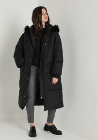 Abrigo puffero largo negro con capucha de piel sintética, botones a presión, dos bolsillos frontales y un diseño acolchado. Llevado sobre un suéter oscuro y jeans gris.