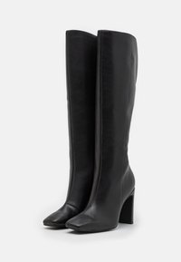 Bottes en cuir noires montant jusqu'au genou, à texture lisse, bout carré et talon haut épais, arborant un design élégant et minimaliste.