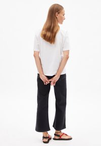 Witte katoenen t-shirt, relaxed fit, korte mouwen. Donkerdenim flared jeans met ruwe randen. Zwarte sandalen met rode accenten. Model met de rug naar de camera.