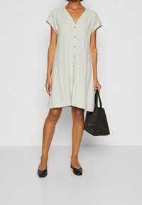 Ljust grön linneliknande klänning med V-ringning, korta ärmar, knapprad fram och en lös silhuett, kombinerad med en svart tote-väska och ballerinaskor.