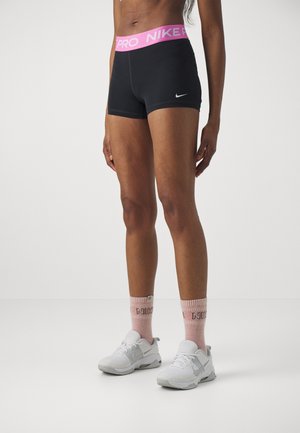 Kobieta nosząca czarne spodenki Nike Pro z różową gumą w pasie, w połączeniu z różowymi skarpetkami Nike i białymi butami Nike, stojąca na białej podłodze.