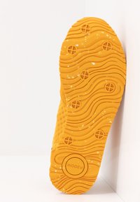 Semelle de chaussure jaune avec un motif de bande de roulement ondulé, des crampons circulaires, de petits débris blancs et le logo "WODEN" embossé près du talon.