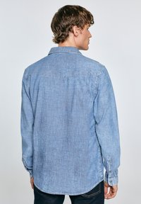 Camisa de mezclilla azul claro con mangas largas, puños abotonados, un solo bolsillo en el pecho y un dobladillo curvado, con una textura sutil.