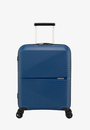 Valigia rigida blu con superficie testurizzata, manico telescopico, quattro ruote e etichetta con logo frontale. Design compatto e rettangolare.