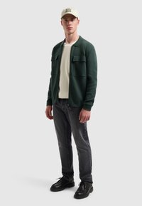 Groene jas met twee voorkruiken, beige shirt eronder, donkere spijkerbroek en zwarte veterschoenen. Heeft een lichtbeige pet.