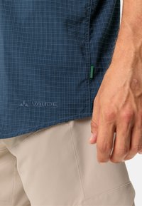 Camisa a cuadros azul marino con una textura sutil, que presenta una etiqueta verde y un logo bordado. Mano mostrada contra pantalones cortos beige claros.