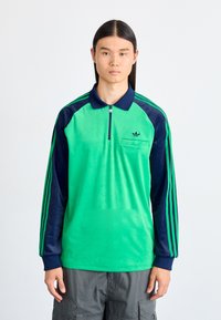 Zelena i tamnoplava polo majica s dugim rukavima, djelomičnim patentnim zatvaračem, džepom na prsima i tri pruge na svakom rukavu. Mekani tekstura, sportski dizajn.