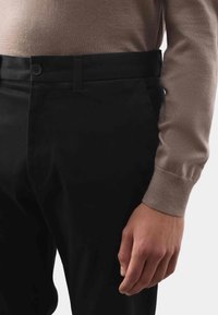 Czarne spodnie o gładkiej fakturze z zapięciem na guzik w pasie, noszone z jasnobrązowym swetrem z dzianiny, częściowo widoczny rękaw i dłoń.