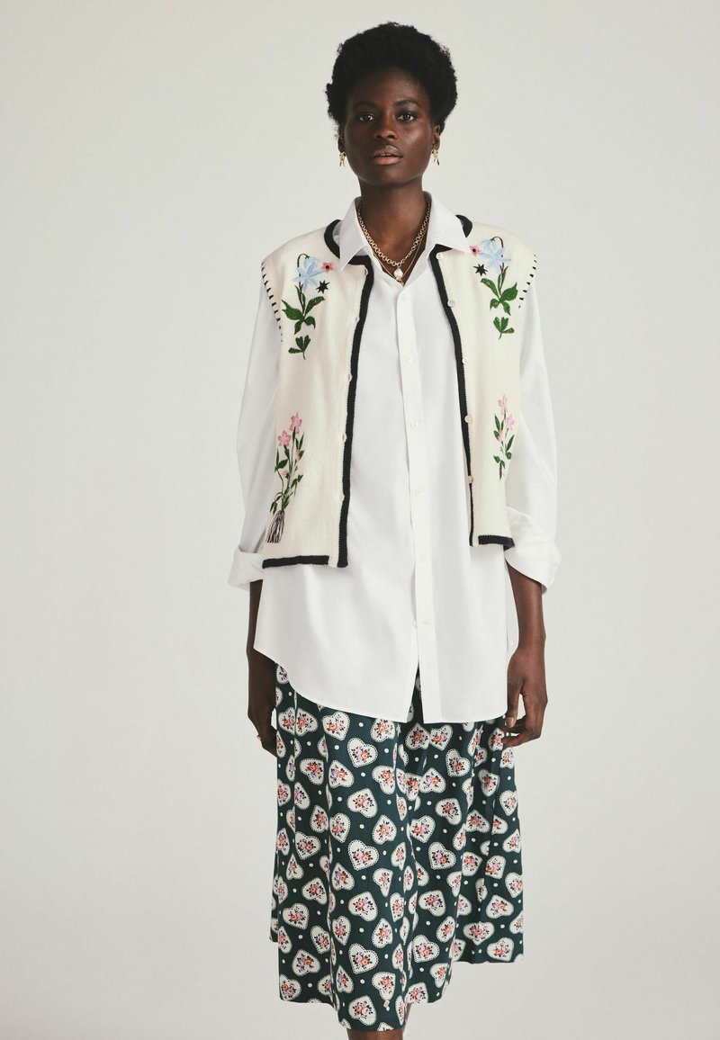 Camisa blanca de manga larga debajo de un chaleco crema con bordados florales; falda verde estampada con motivos rojos y blancos. Textura de tela suave.