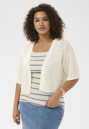 Vrouw met krullend haar die een wit opengewerkt kort mouwig vest, een gestreepte tanktop, een blauwe spijkerbroek en een gouden armband draagt.