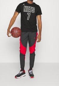Černé tričko s nápisem "NETS" a číslem "7" v bílé barvě, spárované s černými a červenými joggery. Drží hnědý basketbal a má na sobě černé tenisky s červenými akcenty.