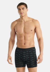 Muž bez trička s krátkými kudrnatými vlasy, oblečený v černých vzorovaných boxerách, stojící na jednobarevném světlém pozadí.