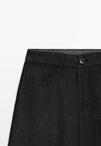 Schwarze Denim-Jeans im Taillenbereich mit Knopfverschluss, Gürtelschlaufen und sichtbarer Vorderseite vor weißem Hintergrund.