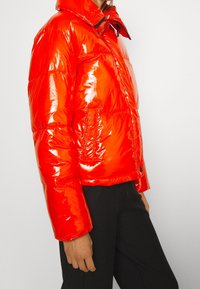 Veste matelassée rouge brillante avec un col haut, des coutures horizontales et une coupe courte. Elle est dotée d'une fermeture par pression et de poches latérales.