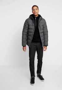 Donzen jack in donkergrijs met een ritssluiting aan de voorkant en een horizontaal gewatteerd patroon. Gecombineerd met een zwarte broek en zwarte sneakers.