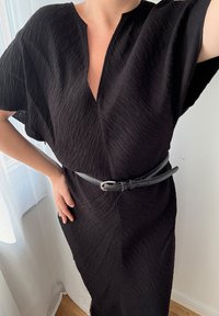 Sort kjole med struktureret stof, korte ærmer og dyb V-hals. Et slankt bælte i sort læder fremhæver taljen. Lys baggrund.