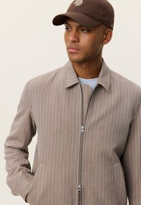 Καφέ ριγωτό μπουφάν με φερμουάρ μπροστά, γιακά και δύο πλαϊνές τσέπες. Φορεμένο πάνω από ένα γαλάζιο πουκάμισο, συμπληρωμένο με καφέ καπέλο.