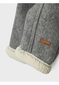 Veste en molleton gris avec un trim en shearling crème, des coutures visibles et une petite étiquette en cuir marron sur la manche.