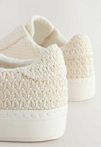 Chaussures à enfiler en toile écrue avec une texture tissée, un talon arrondi et une semelle en caoutchouc blanche. Les détails des coutures le long du bord sont visibles.