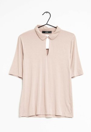 Blouse rose clair à manches courtes avec un col et une ouverture en forme de clé au niveau du cou. Tissu lisse avec une coupe décontractée. Suspendue à un crochet noir.