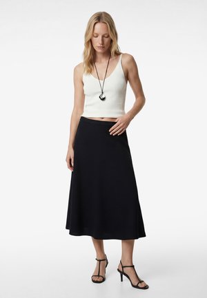 Kobieta ubrana w biały bezrękawnik z krótkim topem, czarną spódnicę midi, czarne sandały na wysokim obcasie i okrągły naszyjnik z zawieszką, stojąca na tle białego tła.