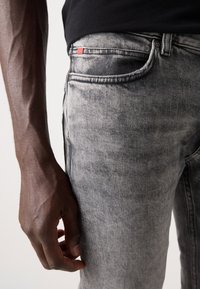 Jeans de denim gris con un ajuste slim, textura desgastada notable y una pequeña etiqueta roja en el bolsillo delantero, mostrados con una mano posicionada cerca.