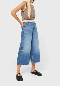 Hellblaue weite Denim-Hosen mit hoher Taille, ausgestattet mit Fronttaschen und einem rohen Saum. Tragen Sie dazu ein braunes, gemustertes Crop-Top und schwarze Slipper.