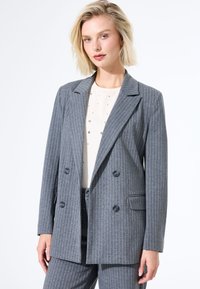 Blazer gris à rayures avec un design croisé, revers crantés et poches latérales. Le tissu semble texturé avec une coupe ajustée.