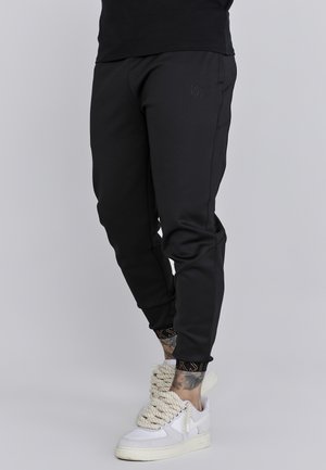 Osoba ubrana w czarne spodnie dresowe i białe tenisówki z grubymi sznurowadłami w stylu liny, stojąca na tle jednolitego jasnoszarego tła.