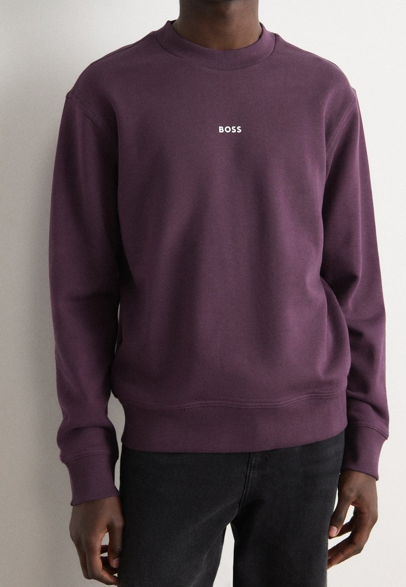 Homme portant un sweat-shirt violet à col rond avec "BOSS" en texte blanc sur la poitrine et un pantalon noir, debout contre un mur clair.
