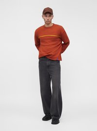 Jeune homme portant une casquette marron, un pull orange avec une bande jaune, un jean noir ample et des chaussures noires, debout devant un fond clair uni.