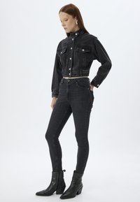 Chaqueta corta de mezclilla negra con cierre de botones, dos bolsillos en el pecho, combinada con jeans ajustados negros y botas de tacón con punta a la altura del tobillo.