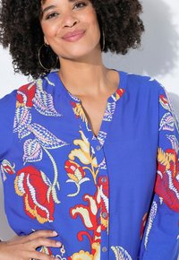 Blaue Bluse mit lebhaften Blumenmustern in Rot, Gelb und Lila. Mit einem Knopfverschluss vorne und einem runden Ausschnitt. Weiche Stoffstruktur.