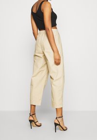 Femme portant un pantalon large cropped beige, un crop top noir et des sandales à talons hauts à lanières noires, se tenant devant un fond blanc uni.