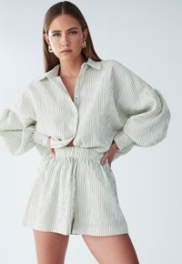 Žena pózujúca s rukami v bok, oblečená v voľnej bielej košeli s zelenými pruhmi a zodpovedajúcich krátkych nohaviciach s vysokým pásom.